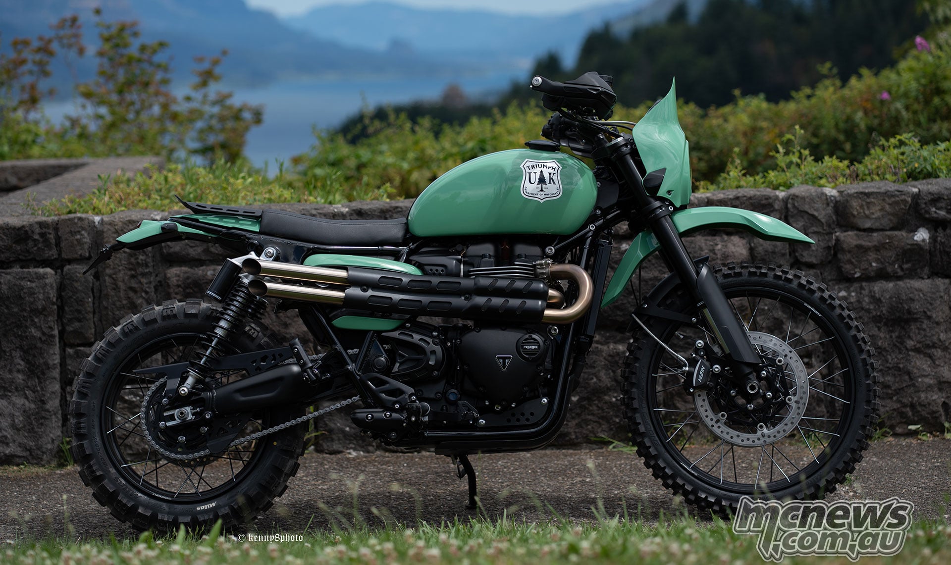 Green Triumph Columbia River Scrambler 1200 X custom with a small screen and rear rack, parked beside a stone wall overlooking mountains and water.