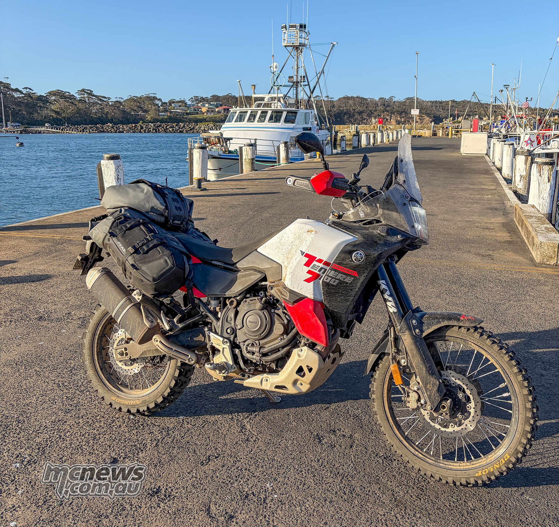 Dust-covered Yamaha Ténéré 700 World Raid with soft luggage parked beside fishing boats at a harbour.
