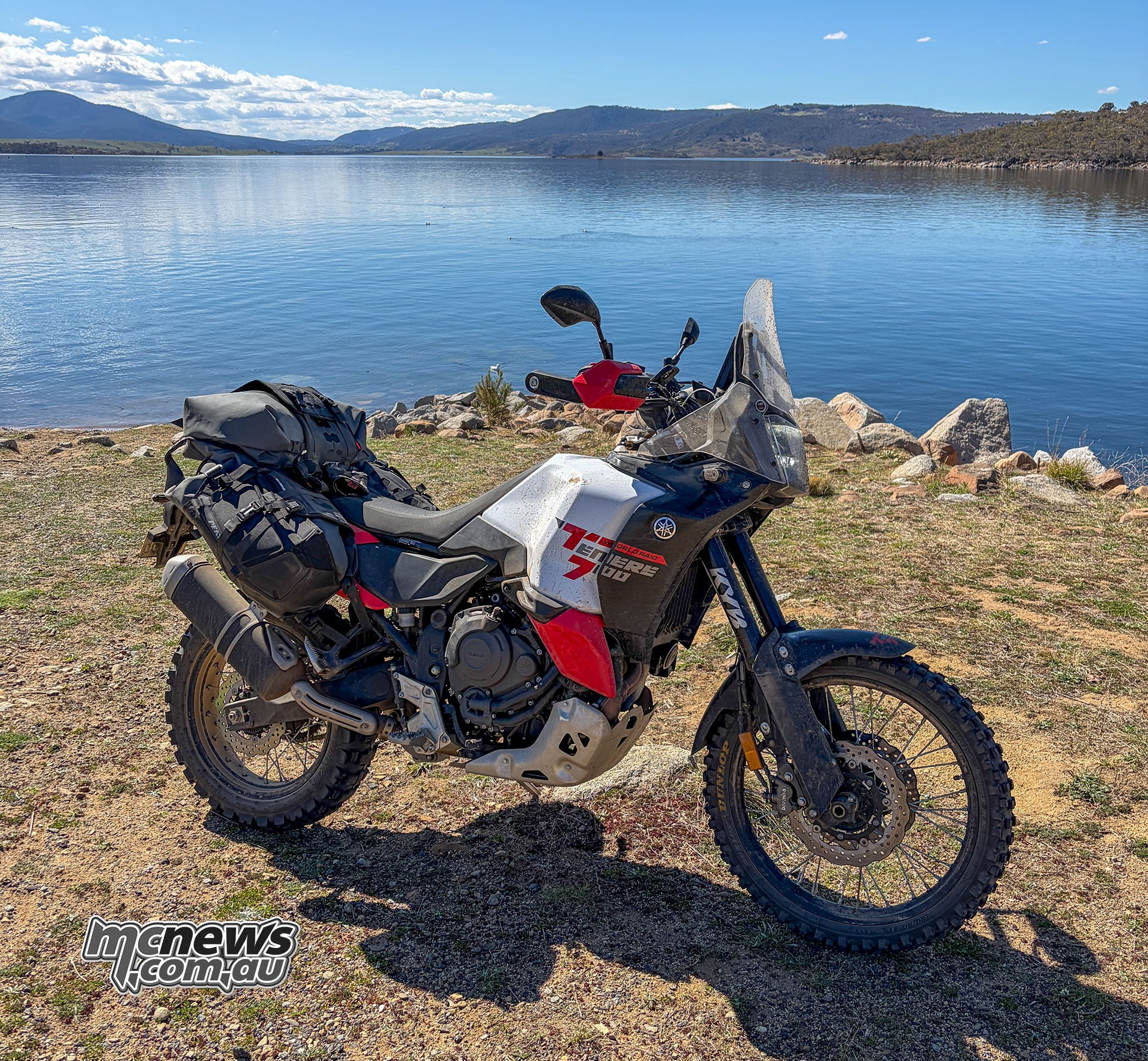 Yamaha Ténéré 700 World Raid carrying soft luggage, parked beside a blue lake with hills on the far shore.