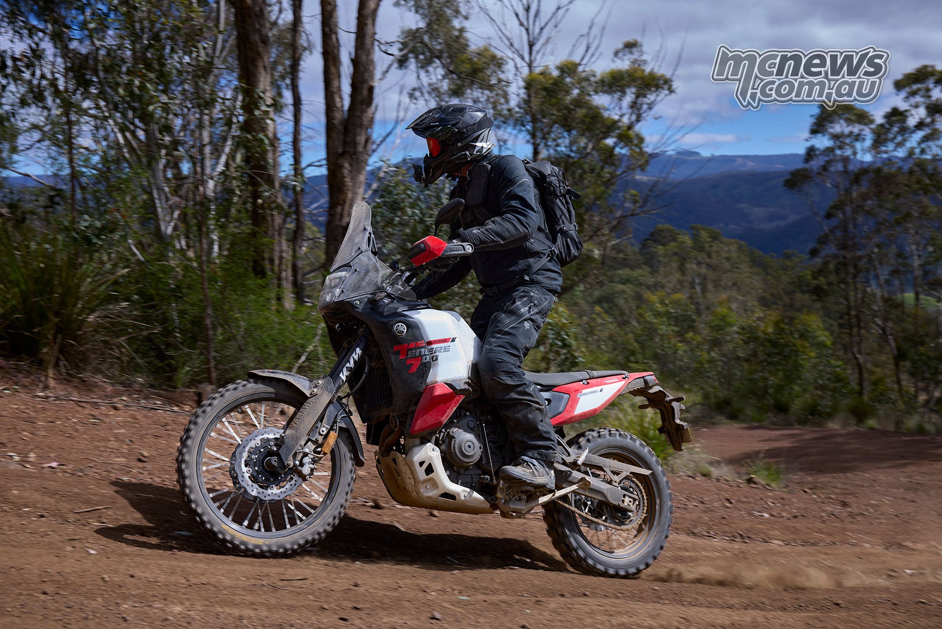Rider standing on a Yamaha Ténéré 700 World Raid while rounding a dirt bend among trees.