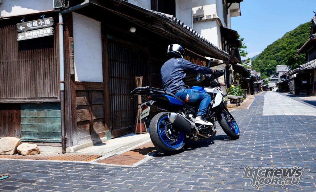 Blue and white Suzuki SV-7GX being ridden slowly through a traditional Japanese streetscape