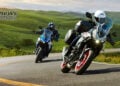 Two 2027 Suzuki SV-7GX motorcycles cornering on a winding road, one blue and white and one Pearl Matte Greige with touring luggage