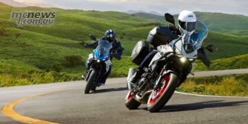 Two 2027 Suzuki SV-7GX motorcycles cornering on a winding road, one blue and white and one Pearl Matte Greige with touring luggage