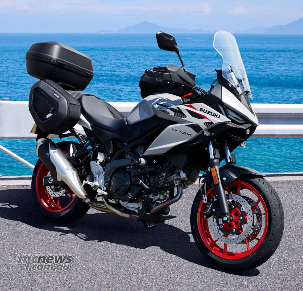 Pearl Matte Greige Suzuki SV-7GX with red wheels, tall windscreen, tank bag, side bags and top box parked beside the sea