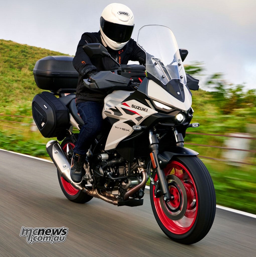 Front three-quarter action view of a Pearl Matte Greige Suzuki SV-7GX with red wheels, tall windscreen and luggage