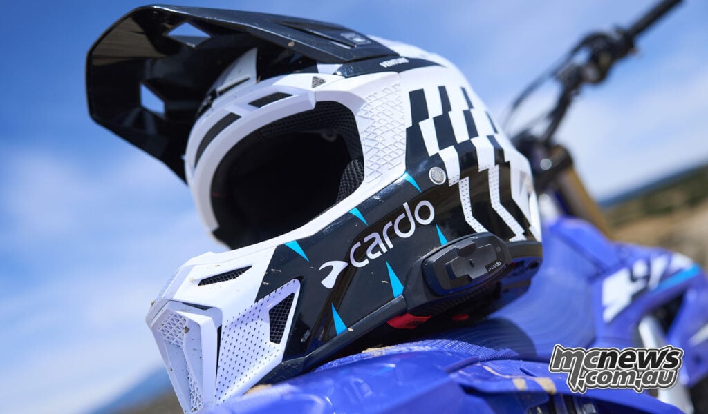 White, black and blue Cardo Venture off-road helmet with an integrated side controller resting on a blue dirt bike.