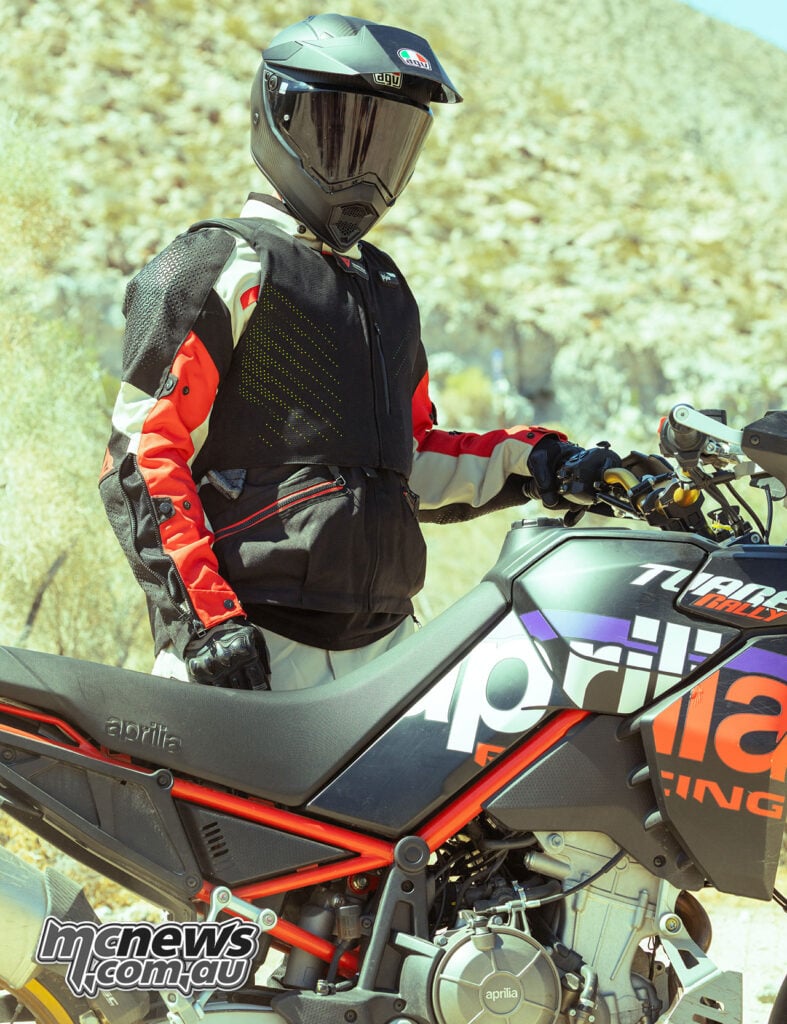 Adventure rider wearing a black Dainese D-air vest while standing beside an Aprilia Tuareg Rally motorcycle in rocky terrain.