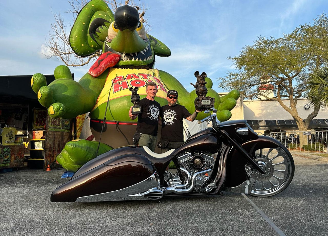 Two men holding Rat’s Hole trophies behind Slow Burn at Daytona, with the event’s giant rat mascot in the background