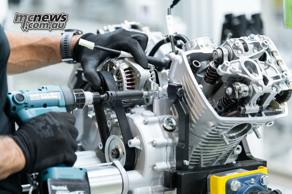Gloved hands secure an alternator on a Moto Guzzi V-twin engine with its rocker cover removed.