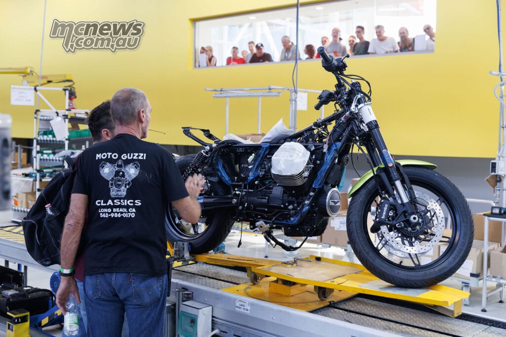 Partly assembled Moto Guzzi on a yellow production carrier, with visitors watching through the window behind.