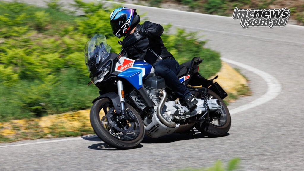 Rennie Scaysbrook leans a blue, white and red Moto Guzzi Stelvio through a bend on a tree-lined road.