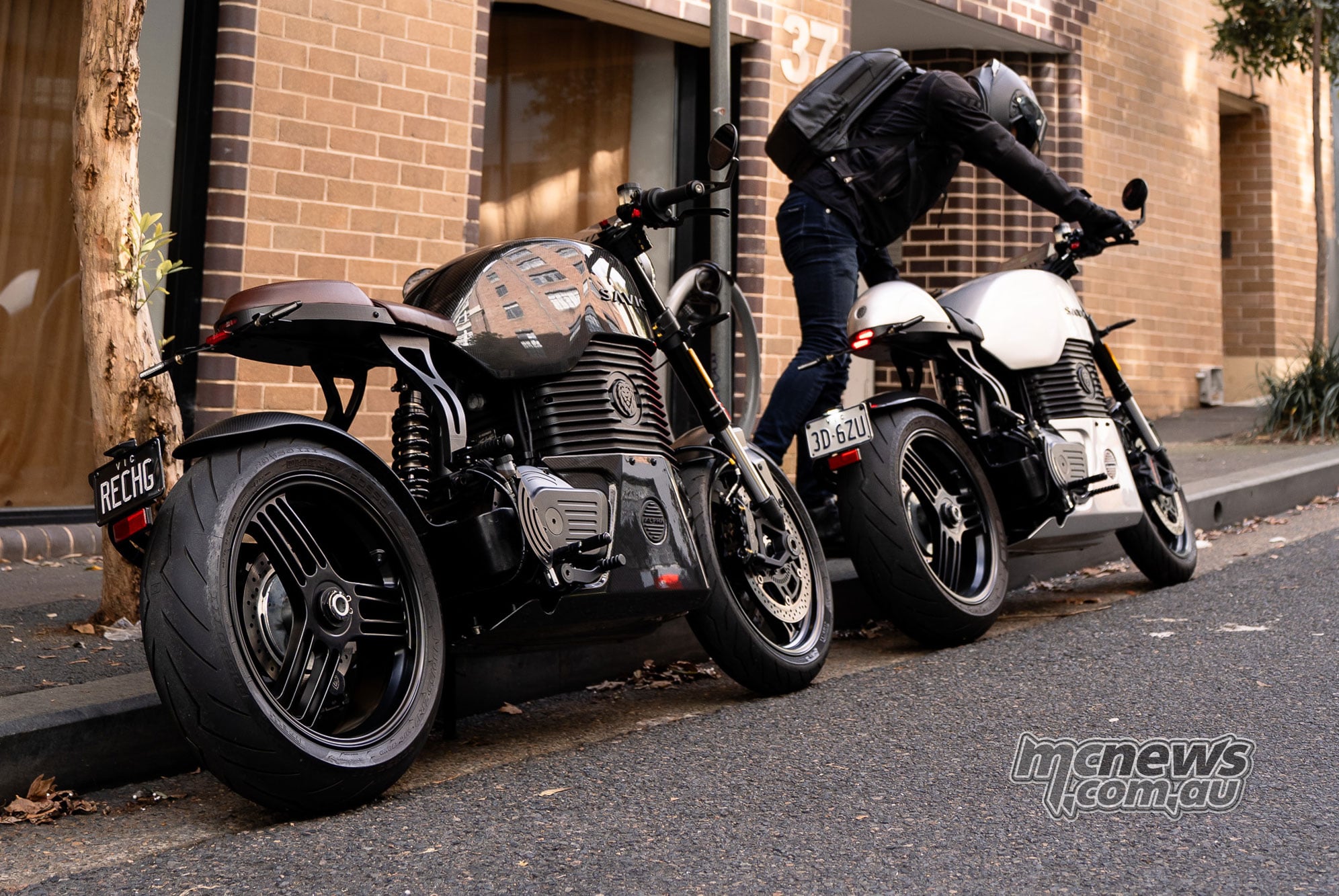 Two Savic C-Series electric motorcycles beside a brick building, with a rider mounting the silver bike.