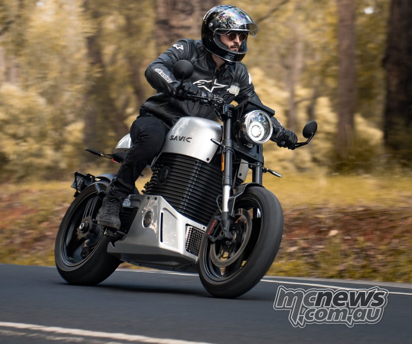 A rider on a silver Savic C-Series electric motorcycle travelling along a tree-lined road.