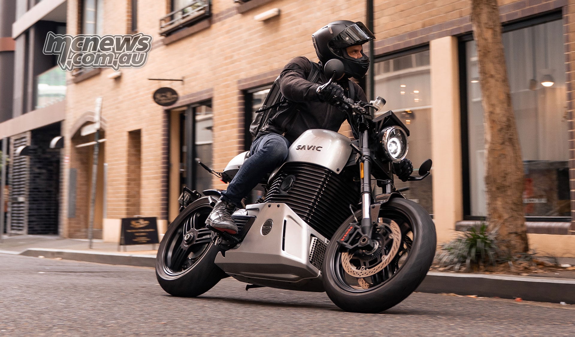 A rider on a silver Savic C-Series electric motorcycle passing brick buildings on an urban street.