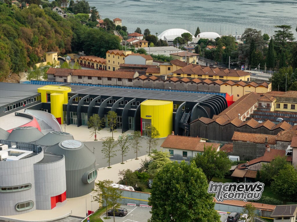 Elevated view of Moto Guzzi’s grey and yellow new buildings among older factory roofs, with Lake Como beyond.