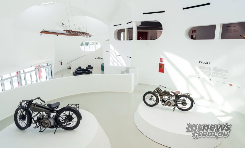 Visitors lean in and take photographs of an early olive-green Moto Guzzi displayed on a white museum plinth.