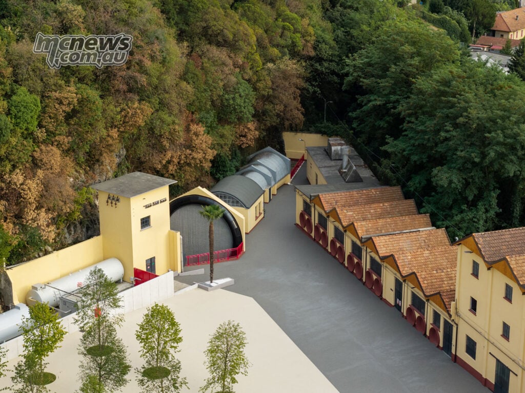 Moto Guzzi’s restored wind tunnel, with its large circular opening beside yellow factory buildings and a wooded hillside.