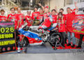 Ryo Mizuno sits on his Ducati Panigale V4 R surrounded by Team Kagayama members, a championship pit board and a celebratory banner.