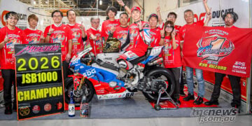 Ryo Mizuno sits on his Ducati Panigale V4 R surrounded by Team Kagayama members, a championship pit board and a celebratory banner.