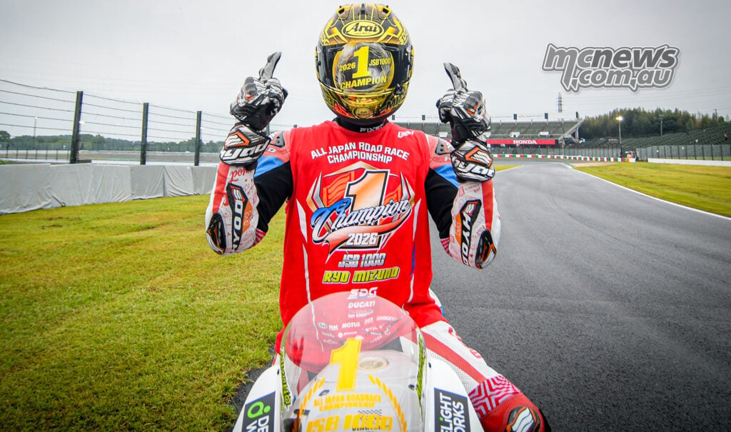 Ryo Mizuno raises both index fingers while seated on his Ducati, wearing a gold champion helmet and red JSB1000 title shirt.