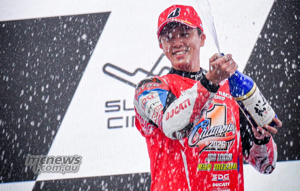 Ryo Mizuno sprays a celebratory drink on the podium while wearing a red cap and his 2026 JSB1000 champion shirt.