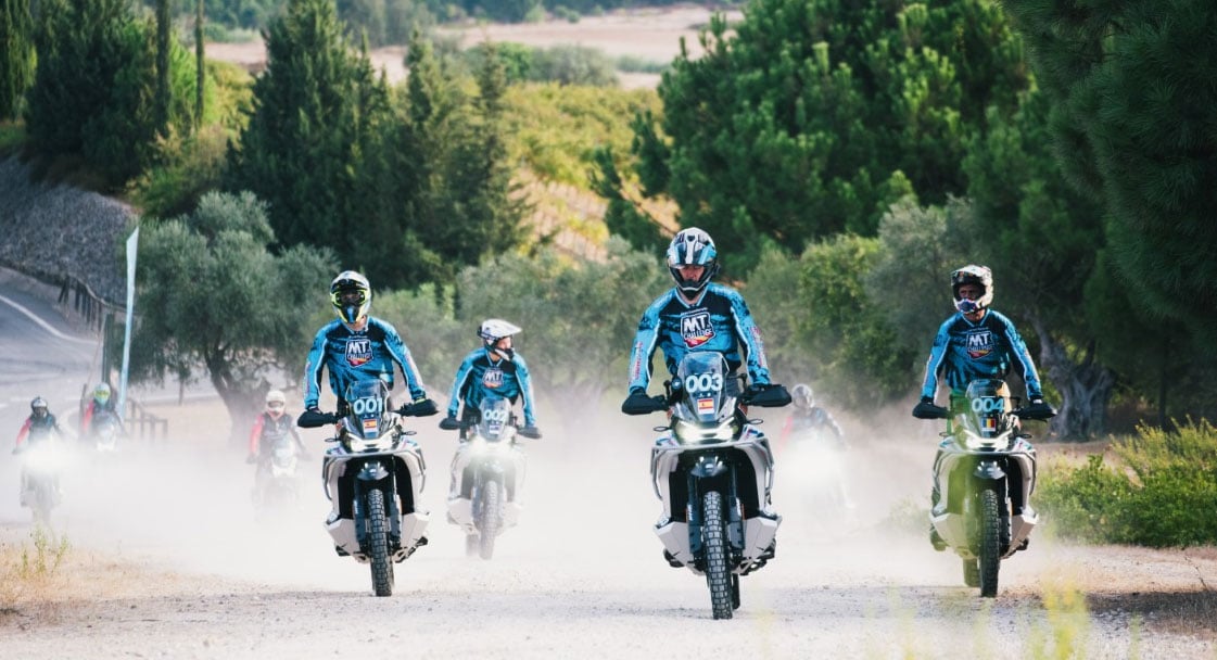 Four adventure motorcyclists riding towards the camera along a dusty unsealed road surrounded by trees.