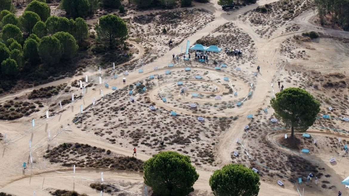 Aerial view of a large circular off-road motorcycle skills course laid out on sandy ground among trees.