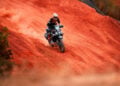 Adventure rider on a CFMOTO 1000MT-X climbing a steep red dirt slope and trailing dust.
