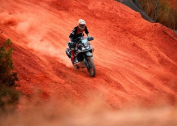 Adventure rider on a CFMOTO 1000MT-X climbing a steep red dirt slope and trailing dust.