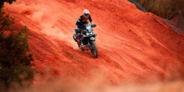 Adventure rider on a CFMOTO 1000MT-X climbing a steep red dirt slope and trailing dust.