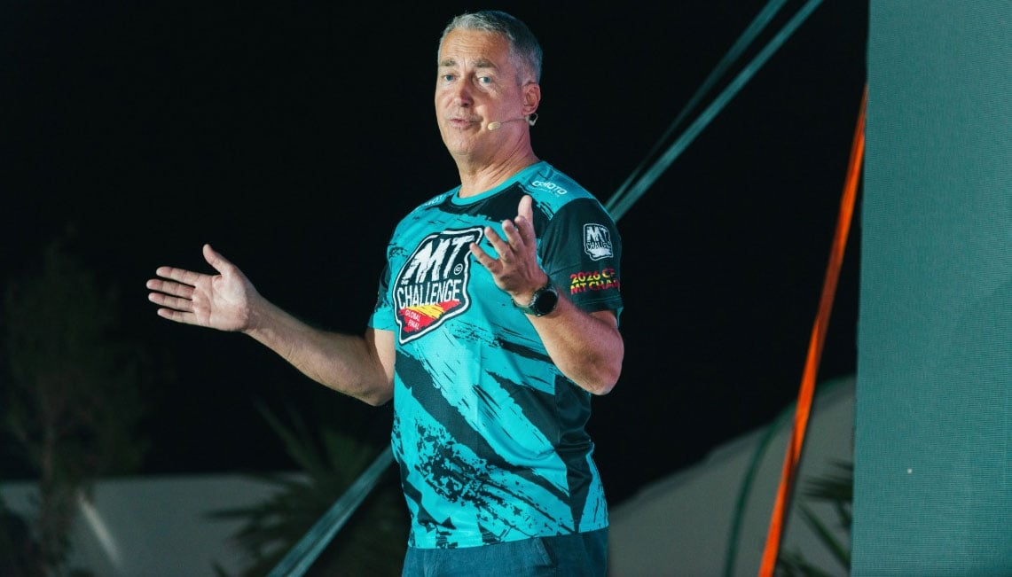 Man wearing a CFMOTO MT Challenge shirt speaking on a stage with a headset microphone.