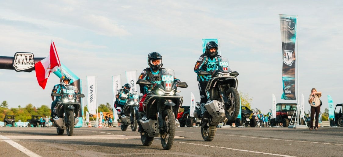 Several riders accelerating away on CFMOTO adventure motorcycles with the nearest rider lifting the front wheel.