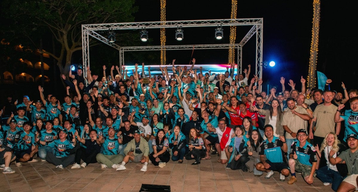 Large group of riders, staff and guests gathered together and cheering at an outdoor evening celebration.