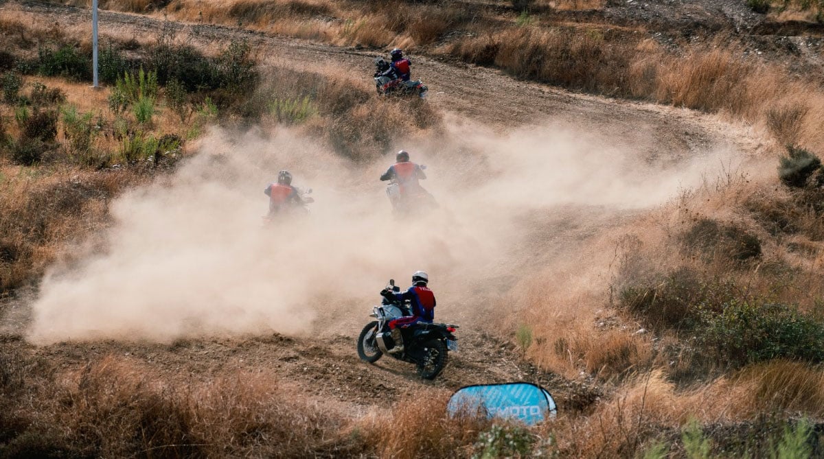 Several adventure riders travelling along a dry off-road trail through a large cloud of dust.