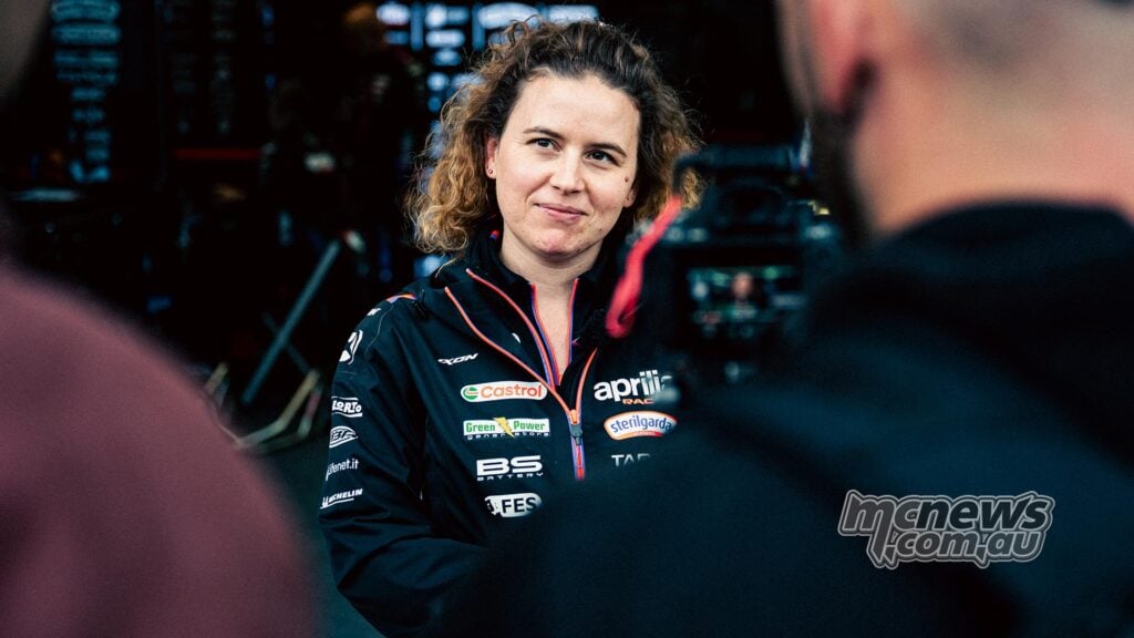 Elena De Cia smiles towards the camera in the Aprilia Racing garage, wearing her black team jacket with other personnel blurred in the foreground.