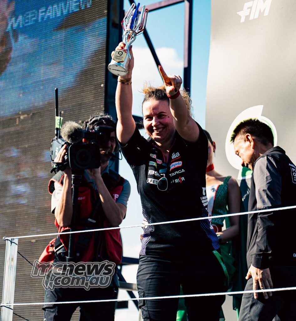 Aprilia Racing's Elena De Cia celebrates on a MotoGP podium, holding a trophy above her head and raising one finger, with a television cameraman alongside the stage.