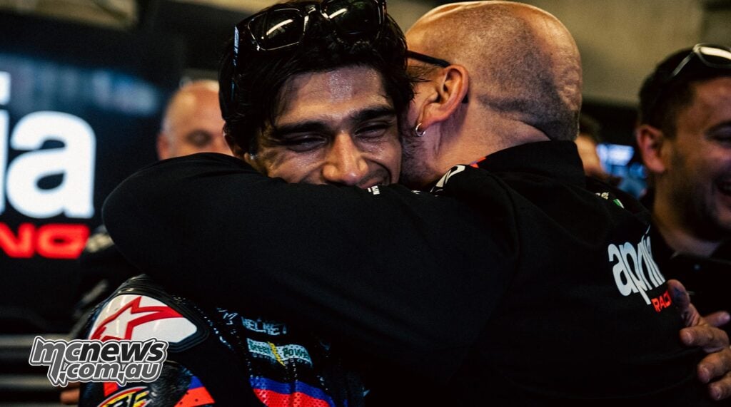 Fabiano Sterlacchini embraces a smiling Aprilia MotoGP rider wearing black racing leathers with orange and blue detailing.