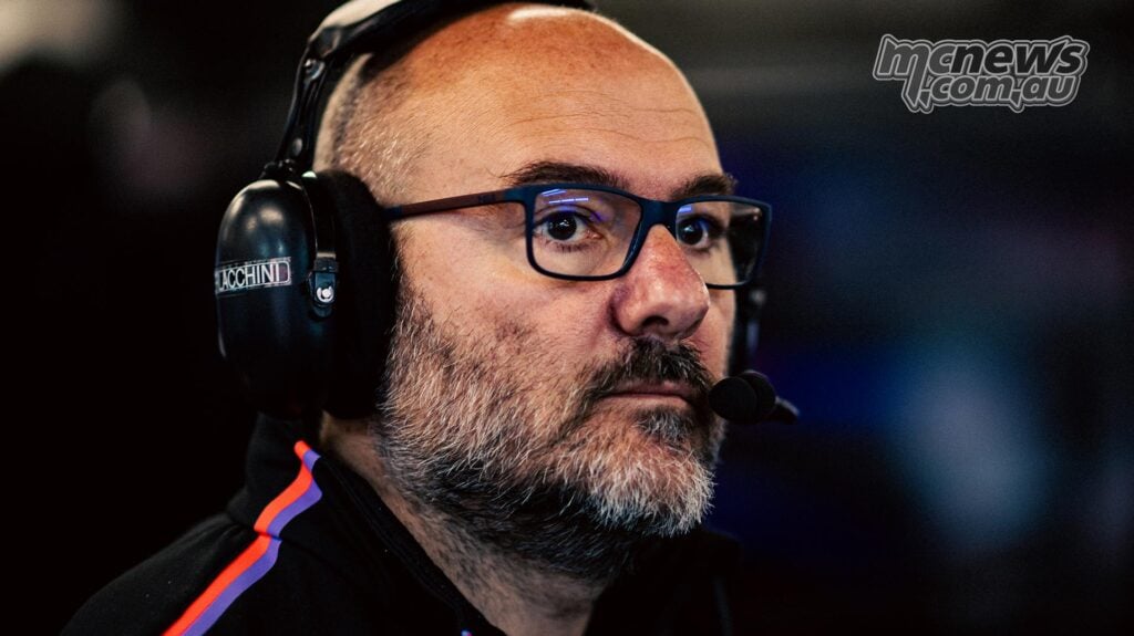 Close-up of Fabiano Sterlacchini wearing blue-framed glasses and a team headset, concentrating on activity away from the camera inside the Aprilia MotoGP garage.