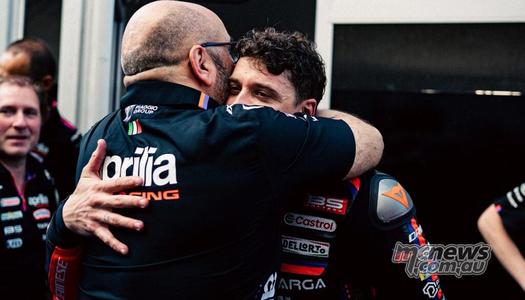 Fabiano Sterlacchini embraces an Aprilia Racing rider in black, orange and blue leathers, with the rider looking over his shoulder and other team members nearby.