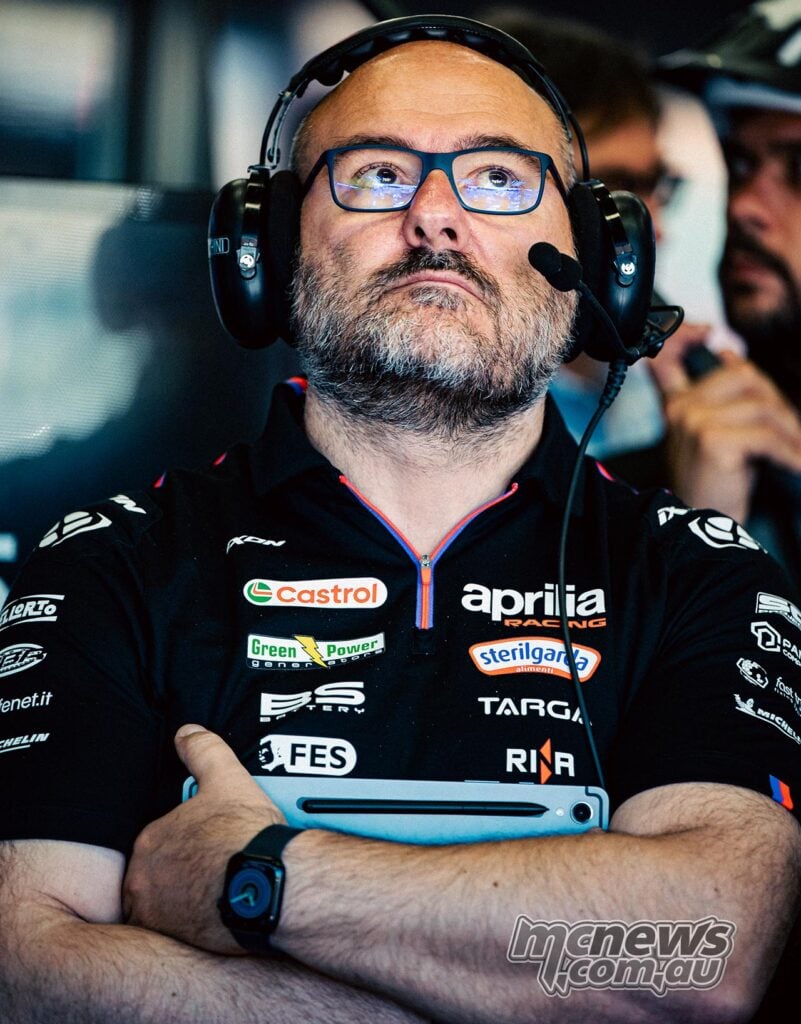 Fabiano Sterlacchini watches overhead monitors in the Aprilia Racing garage, wearing a headset with his arms folded and a blue tablet tucked beneath one arm.