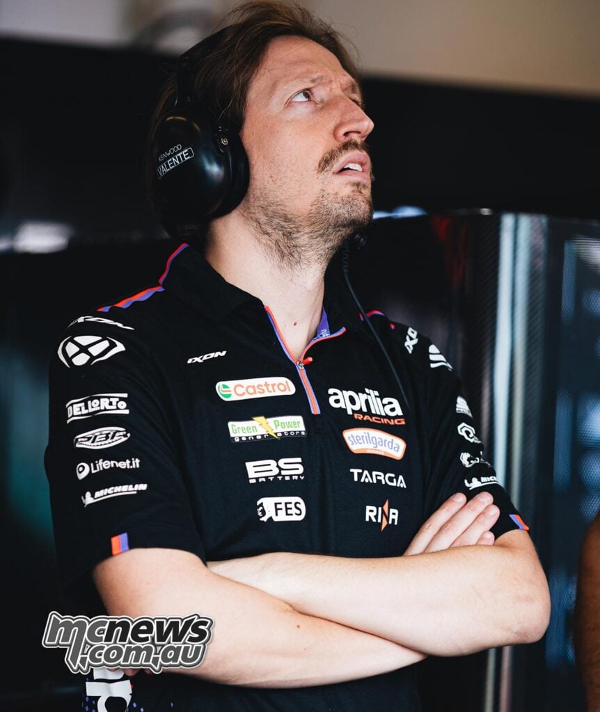Aprilia Racing mechanical engineer Pasquale Valente stands with his arms folded in the MotoGP garage, wearing a communications headset and looking upwards towards off-camera monitors.