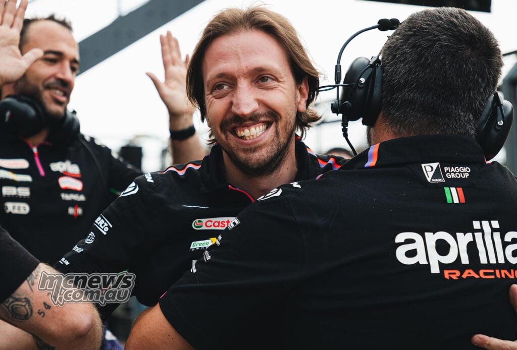 Pasquale Valente smiles while interacting with fellow Aprilia Racing team members in the MotoGP paddock, with a colleague wearing a headset in the foreground.