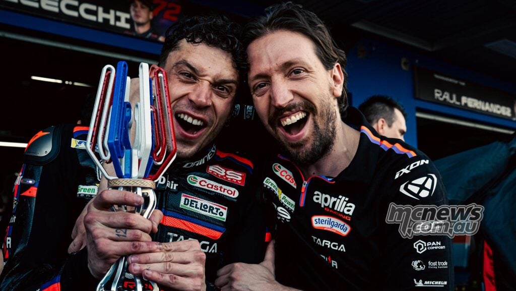 Aprilia Racing engineer Pasquale Valente celebrates alongside Marco Bezzecchi, who is smiling and holding a racing trophy in his Aprilia MotoGP leathers.