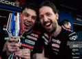 Aprilia Racing engineer Pasquale Valente celebrates alongside Marco Bezzecchi, who is smiling and holding a racing trophy in his Aprilia MotoGP leathers.
