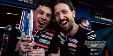 Aprilia Racing engineer Pasquale Valente celebrates alongside Marco Bezzecchi, who is smiling and holding a racing trophy in his Aprilia MotoGP leathers.