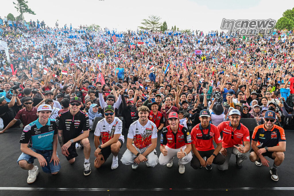 Eight riders crouch at the front of a large crowd waving flags, raising their hands and taking photographs.