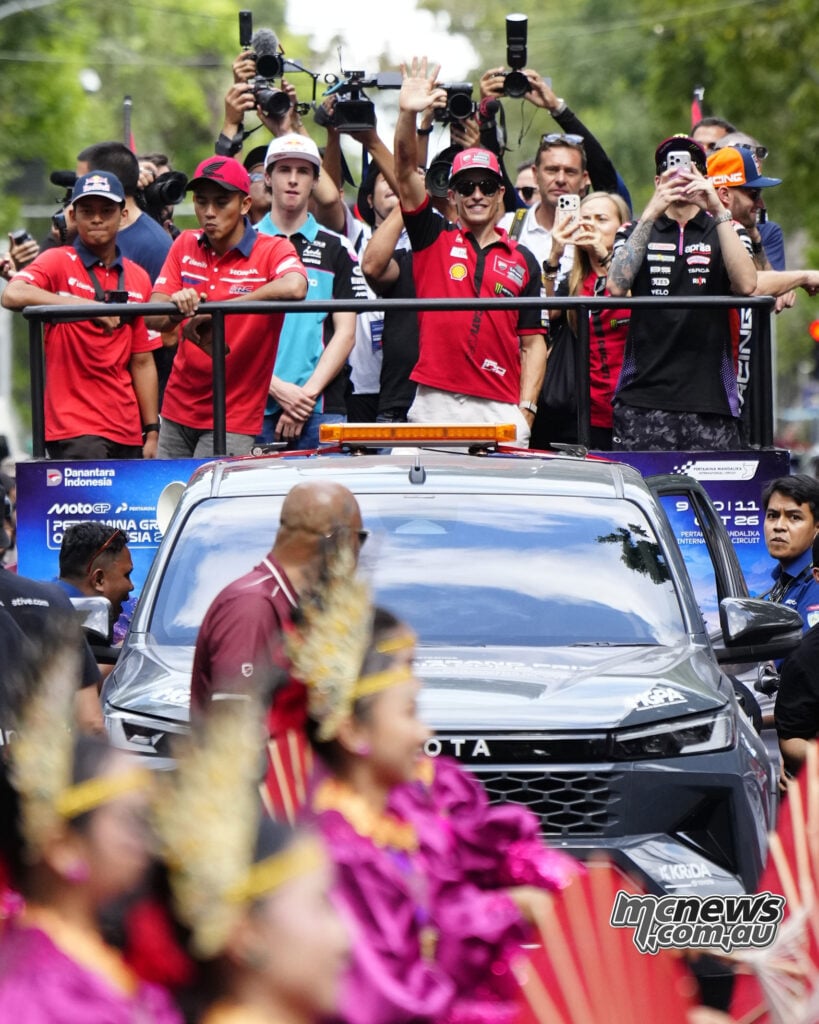 Marc Marquez waves from a ute carrying riders and photographers, with costumed performers in the foreground.