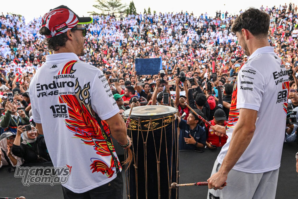 Two VR46 riders in white shirts hold drumsticks around a large decorated drum, facing a packed crowd of fans.