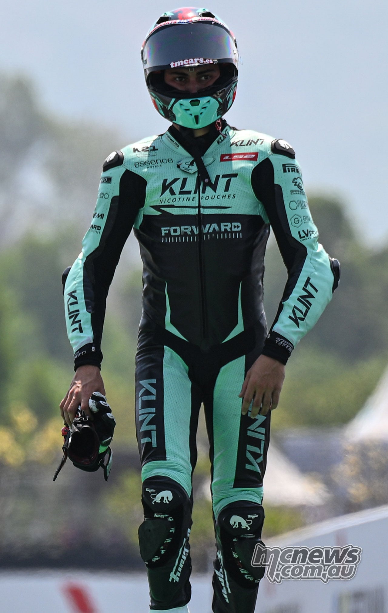 Alex Escrig walks beside the circuit in his turquoise and black racing leathers, carrying his gloves.