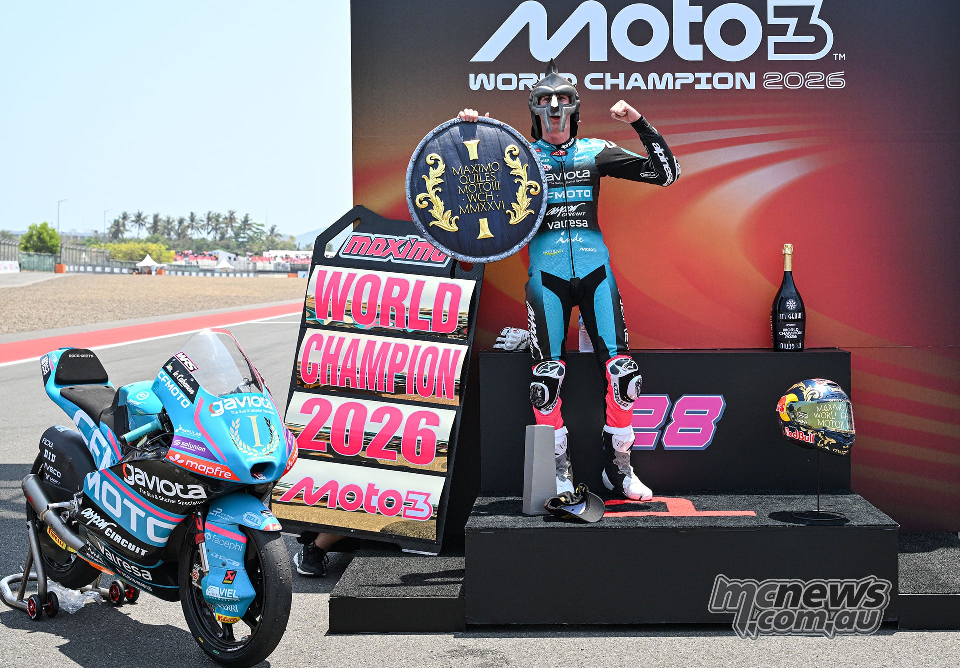 Maximo Quiles wears a metallic mask and holds a shield beside his motorcycle and a 2026 world champion display.